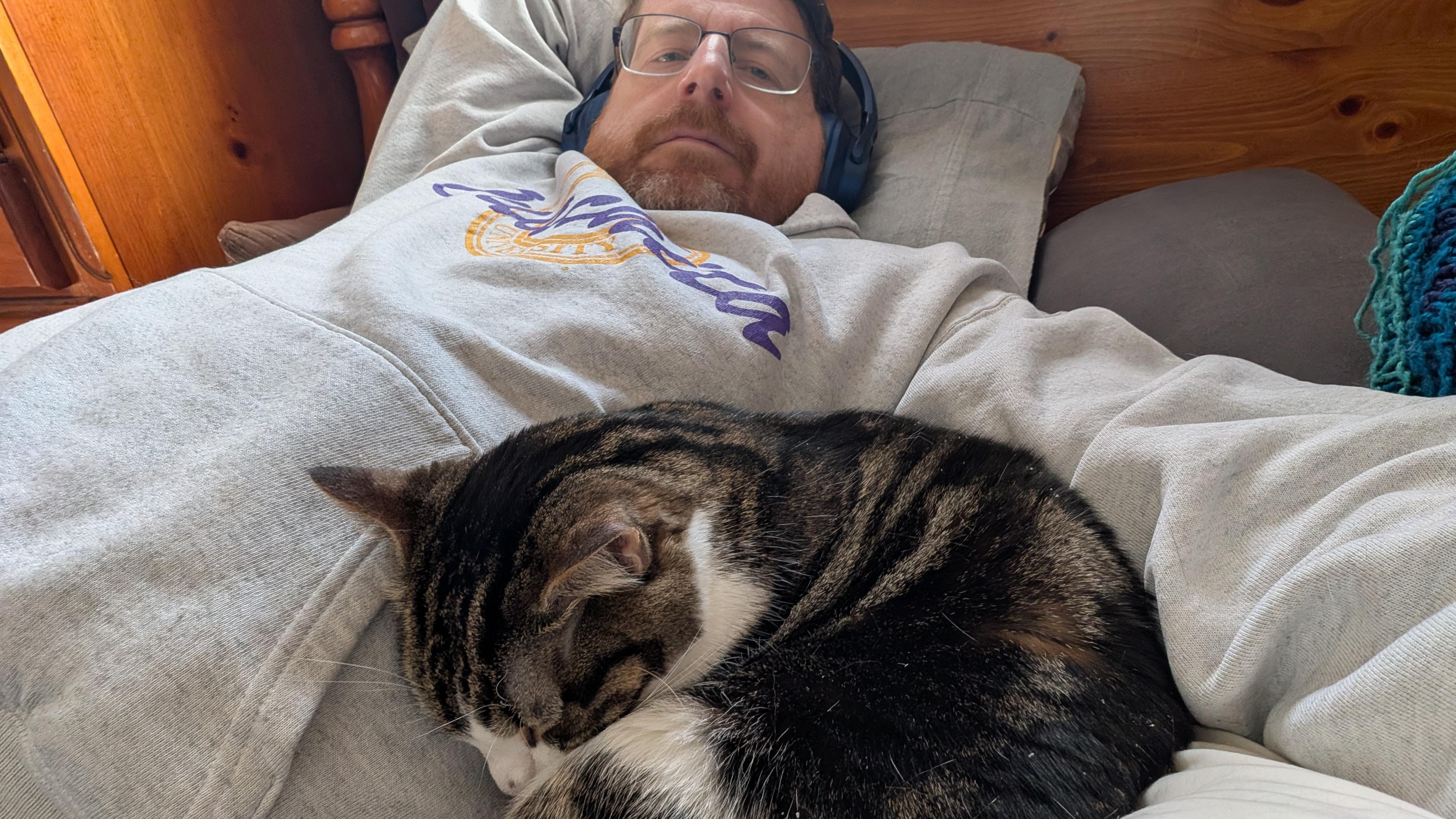 man in a gray hoodie lays on a bed with a brown and white tabby cat curled up into the crook of his left armpit.