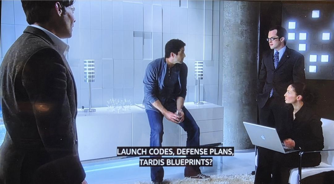 James Callis (standing) as Doctor Grant, Zane and Lupo seated (she's got a laptop open on her lap), and Fargo standing. They are all in Fargo's super modern white office (everything's white in it). They are discussing what Zane found while hacking the DOD. Behind Fargo is a wall with lit up white tiles.