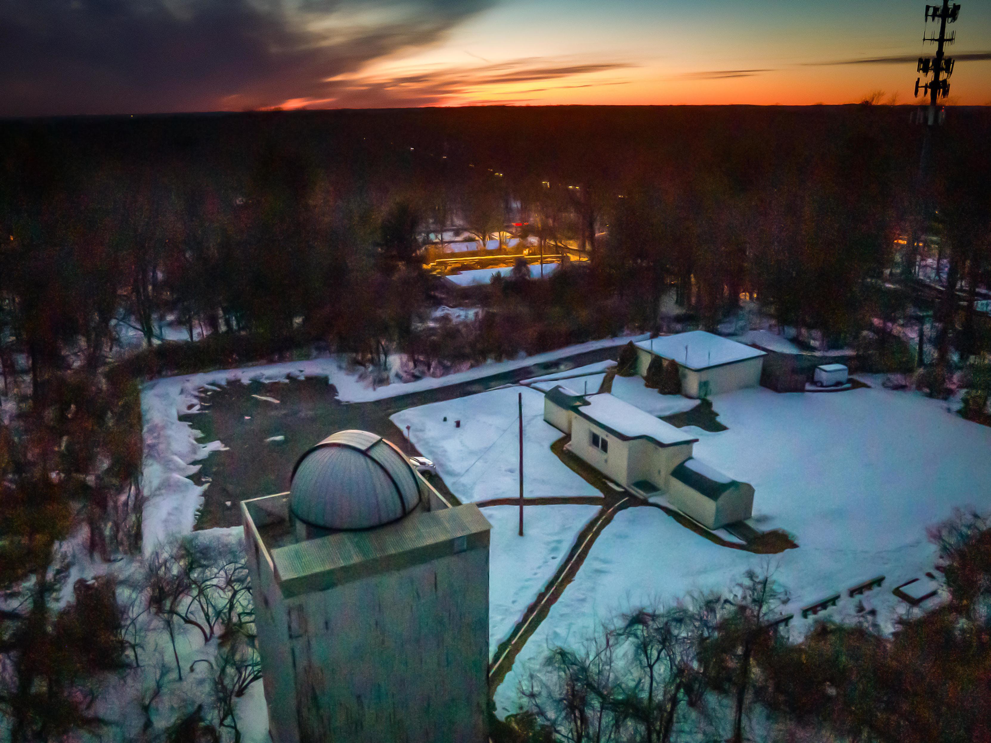 A snowy observatory complex with a dome telescope and adjacent buildings at dusk, surrounded by bare trees and a darkening sky.