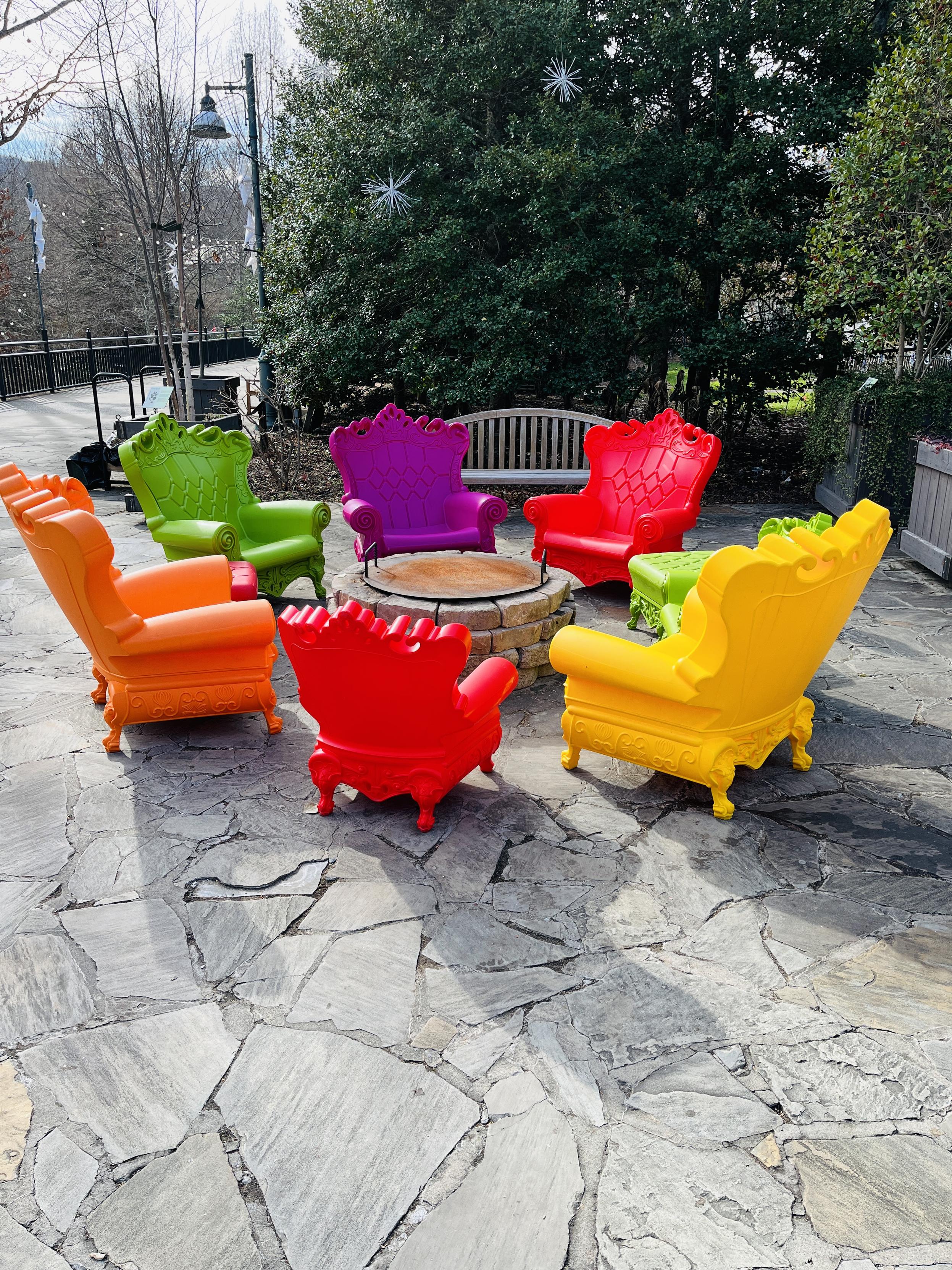 A daytime photo of a ring of very large, bright plastic chairs, in shapes like upholstered furniture sit on a grey surface, surrounding a (non-lit) firepit.  At night, during these events, the fire would be going and the chairs would be very popular.

There are 7 chairs, two probably kiddy-sized, with Queen Anne legs, and rolled arms, and details that make each one look like something from your grandmother's living room, perhaps.  I think there's an ottoman tucked in to that circle as well.

They're in orange, green, yellow, red, and green and such a jolt of color in the winter setting, where all the trees are bare, and it is quite cold.