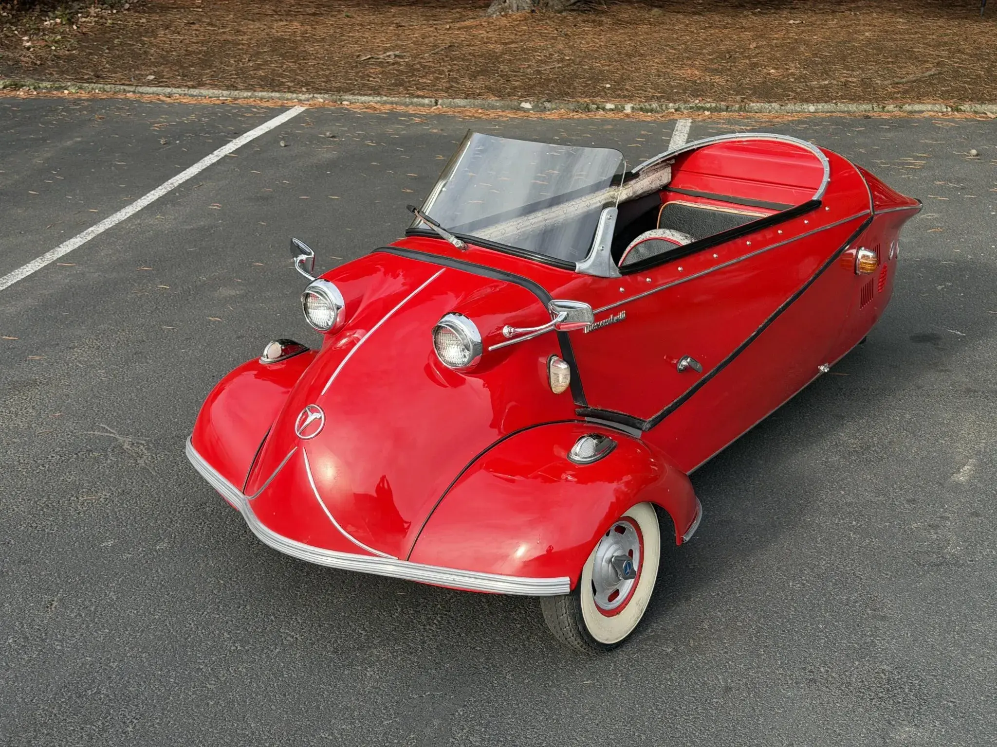 This 1958 Messerschmitt KR201 is powered by a 191cc two-stroke single paired with a sequential four-speed manual transmission and is finished in red over black vinyl with snakeskin trim and red piping. Equipment includes 10″ steel wheels with bright covers, a black soft top, side-mounted turn signals, a steering yoke, tandem seats, and an enclosed drive chain.