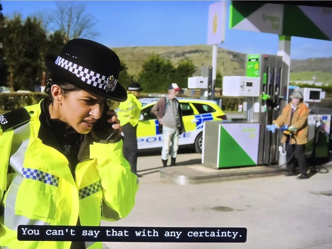 Mandip Gol wearing a constable's uniform with neon yellow jacket and black/white checkered strips and reflective strips. She's on a mobile phone at a gas station. She says "You can't say that with any certainty." Behind her is a police car, civilians and another constable.