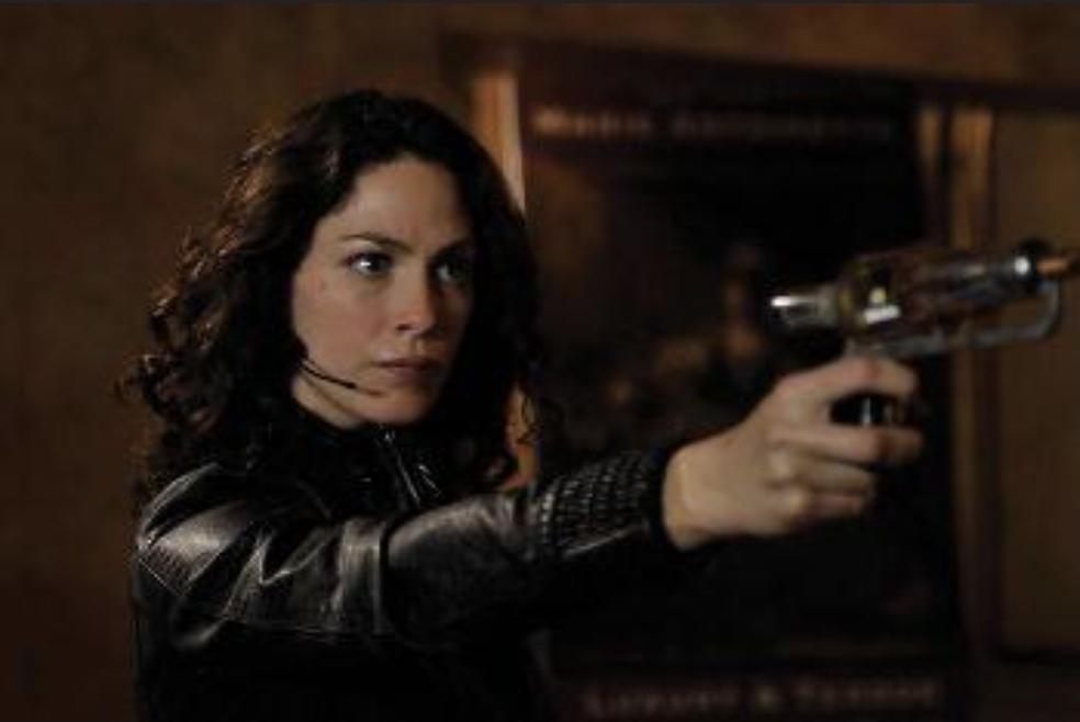 Myka pointing the laser gun that shocks people temporarily. She looks serious and is wearing a black leather jacket and headset. There's a mirror behind her.