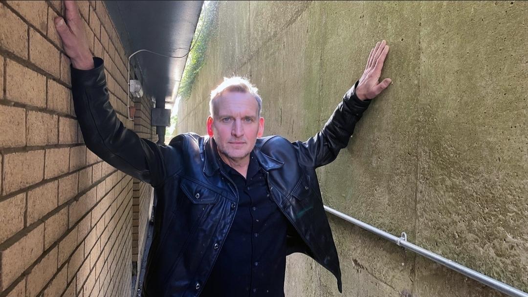 Current photo of Christopher Eccleston in a black jacket and black shirt, standing in a narrow hallway hands resting in both walls above his head as he looks directly at the camera.