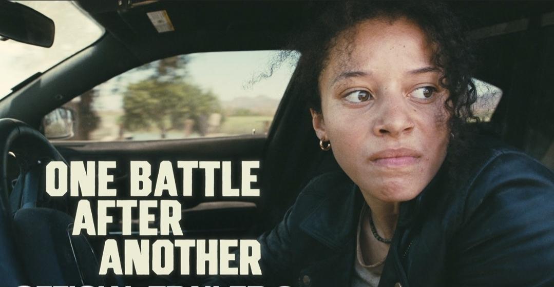 Chase Infiniti as Willa in One Battle After Another. She's looking grim. In a car behind the wheel. Trees outside.