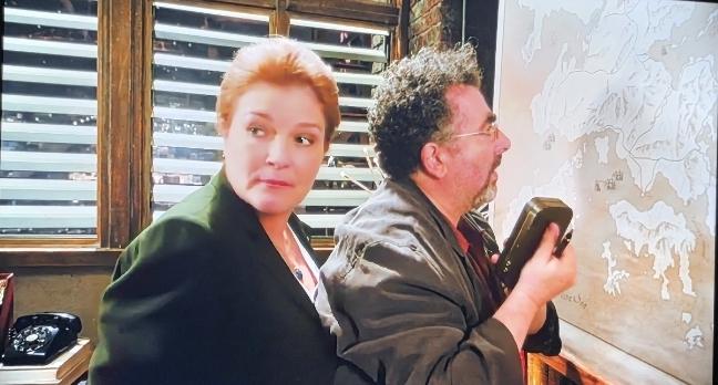 Kate Mulgrew as Jane Lattimer and Artie in the Warehouse in front of a map. Jane is looking over her shoulder. A window with Venetian blinds behind her.