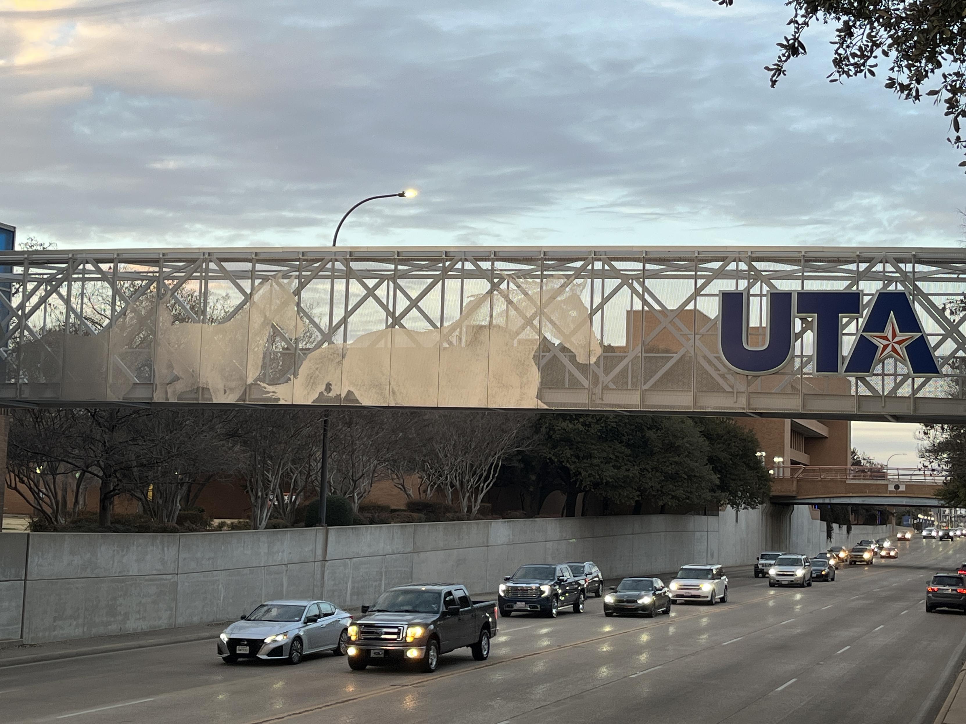 A caged pedestrian bridge over a roadway. Cars on the road have their lights on. The bridge is decorated with metallic horse silhouettes, and the U.T.A. logo.