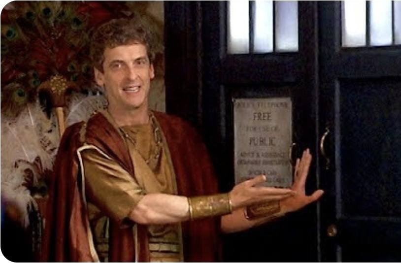Peter Capaldi as a merchant in Pompeii. He has his left hand on the TARDIS and his right hand is pointing at it. He is looking slightly over his shoulder and smiling.
