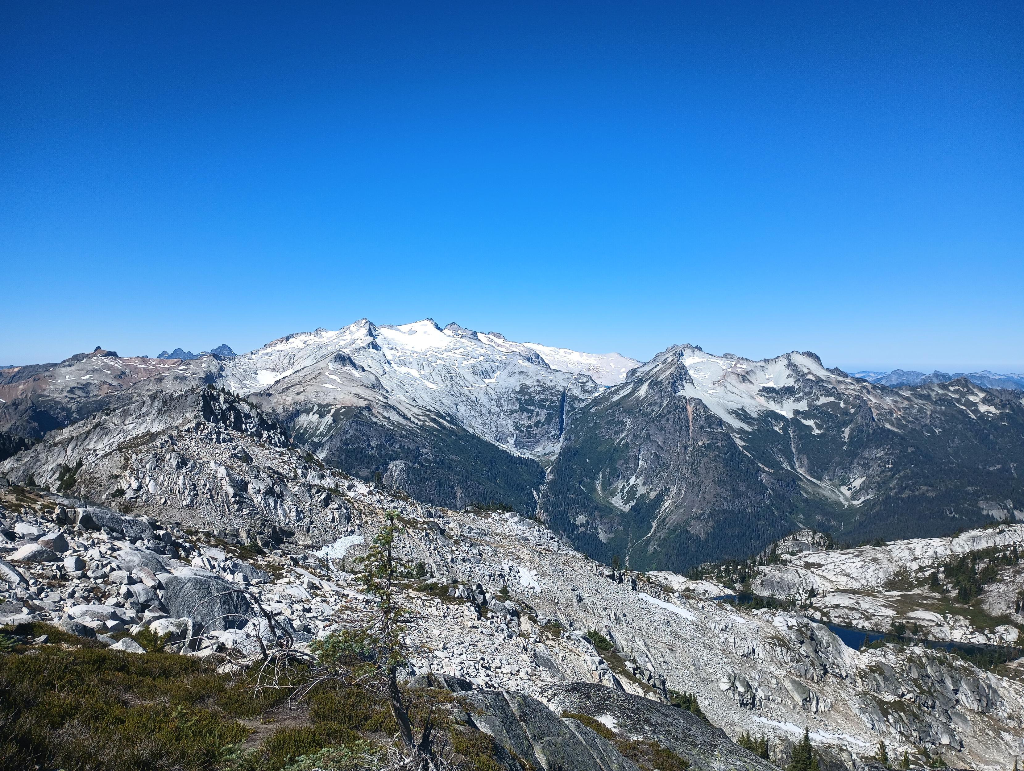 A massive sprawling mountain of light stone and glaciers, its sides carved by waterfalls. As seen from a mountaintop, across the valley, on a bluebird day in summer.