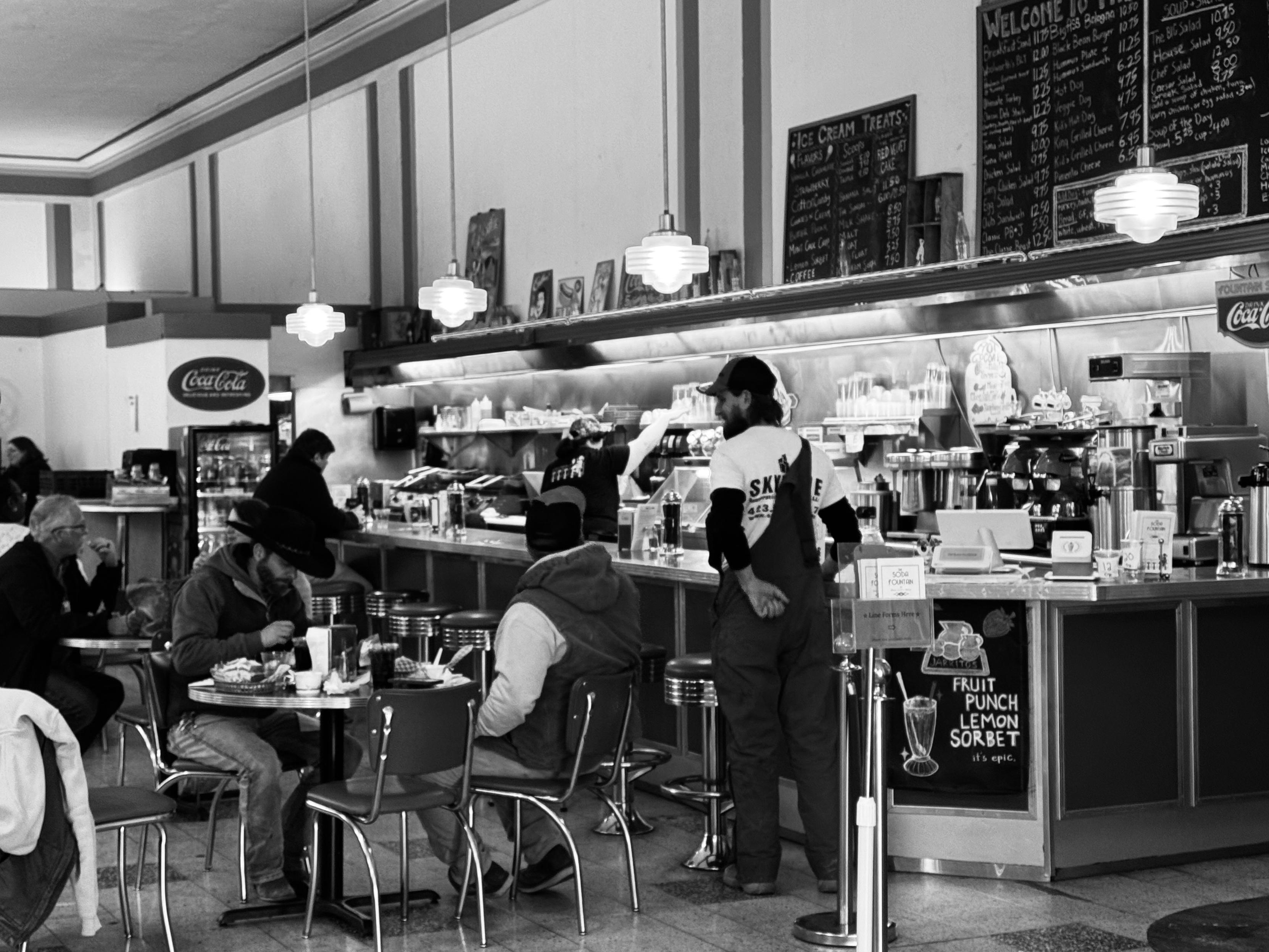 A black and white image of an old-timey lunch counter inside a building now turned into small artists cubes which is a great place to catch lunch or find a painting.  

Many people are sitting at aluminum tables, and a man is standing up near them.  The counter has chrome barstools, and someone is working behind the counter.  The light fixtures are of the era as well.

It would have been a better picture if I'd taken more time, or had Bob Horowitz with me.  I'm always leery of photographing people in the wild...