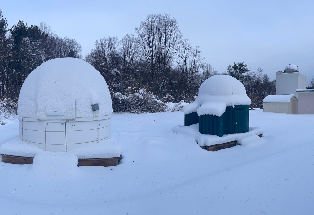 Snow-covered observatory domes and equipment sit in a quiet, snowy field surrounded by bare trees under a clear sky.