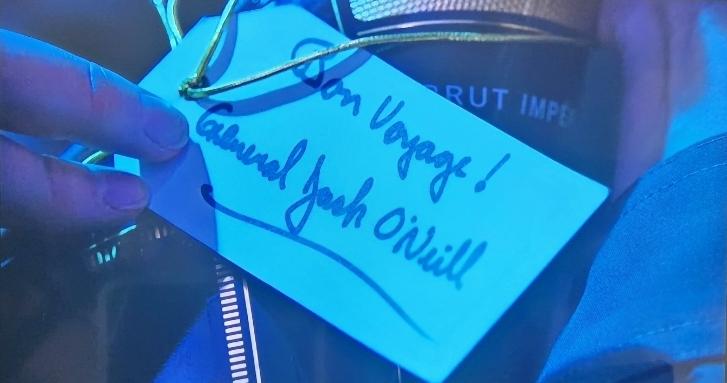 A hand holding a champagne bottle. The gift tag reads "Bon Voyage! General Jack O'Neill"