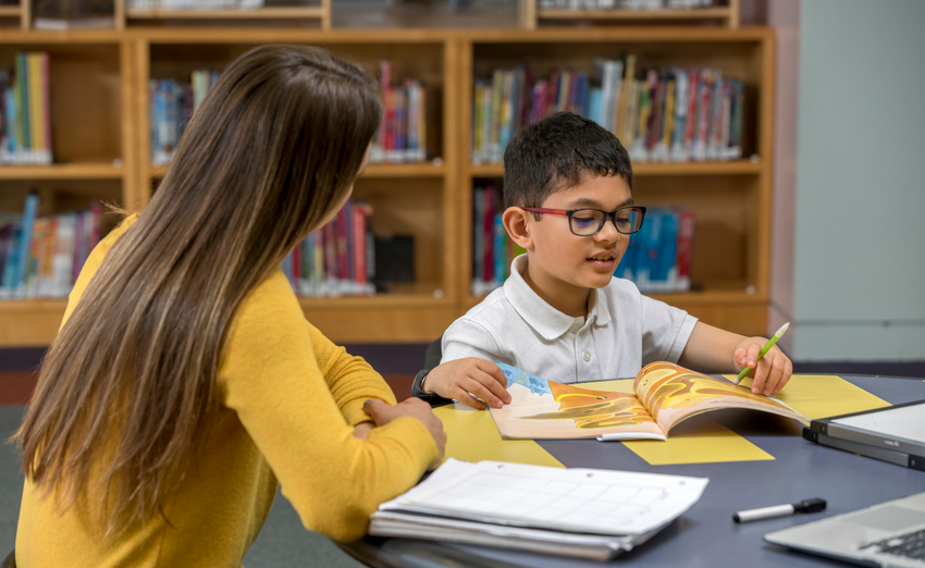 Teacher in a yellow sweater helping a young boy read a colorful book in a school library setting.