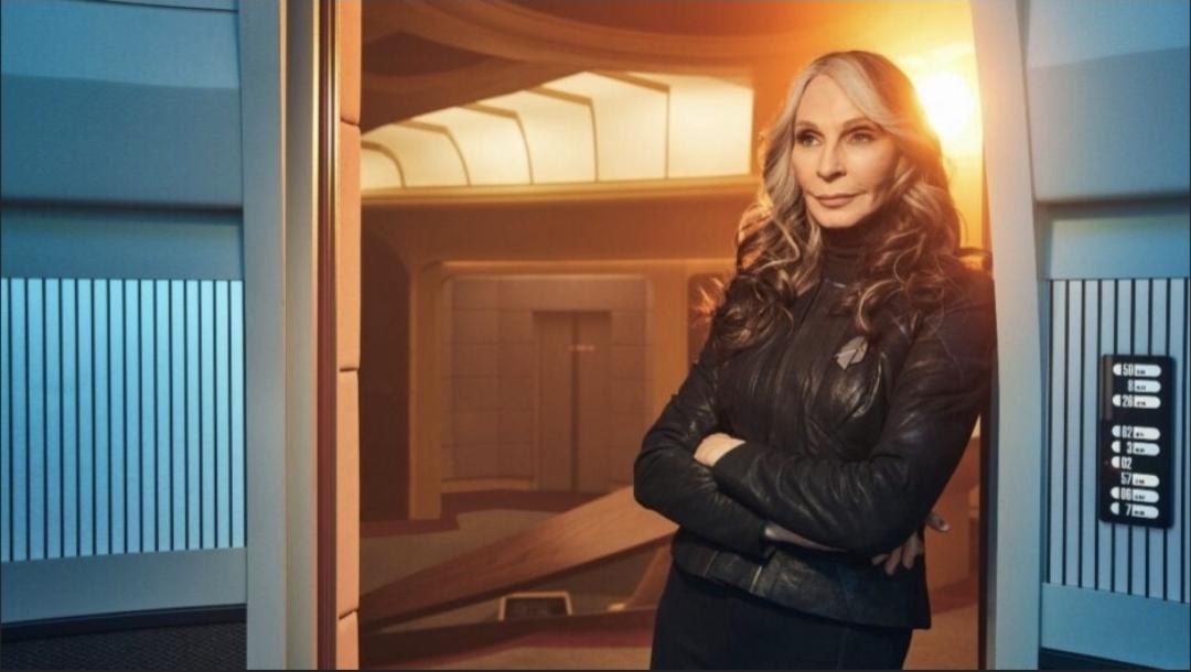 Gates McFadden in Star Trek Picard arms crossed leaning against the blue bulkhead. The bridge is seen behind her. Maybe that's the elevator? There is a golden light behind her. She is wearing a short back leather/ette jacket, black turtleneck and black pants. She is smiling. Maybe sardonically.
