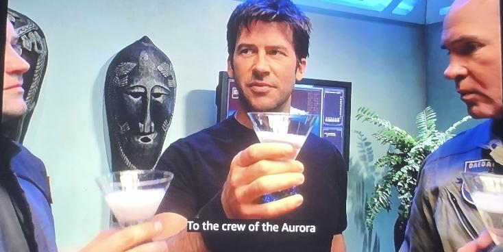 Sheppard, McKay, and Caldwell are shown raising a glass of champagne To the crew of the Aurora. An African mask and computer screen are shown behind them.