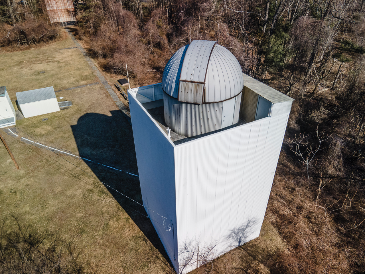 Aerial view of a white observatory tower with a domed roof surrounded by dry trees and grassy fields.
