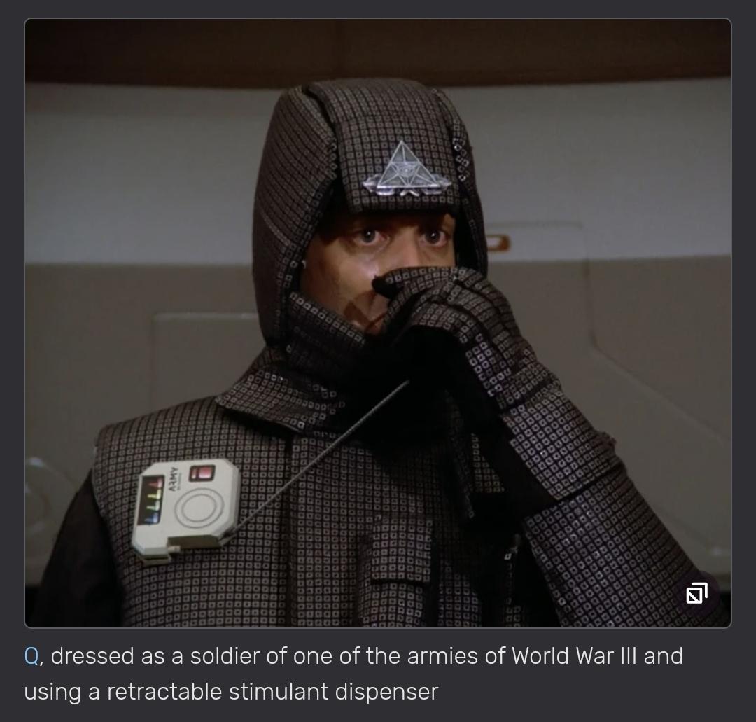 Q is dressed as a soldier of one of the armies of World War III and using a retractable stimulant dispenser. His uniform is dark grey and padded. It includes a hoodie (covering most of the face) and gloves. He is standing in front of a painted wall. It is mostly ecru but with a white band on top.