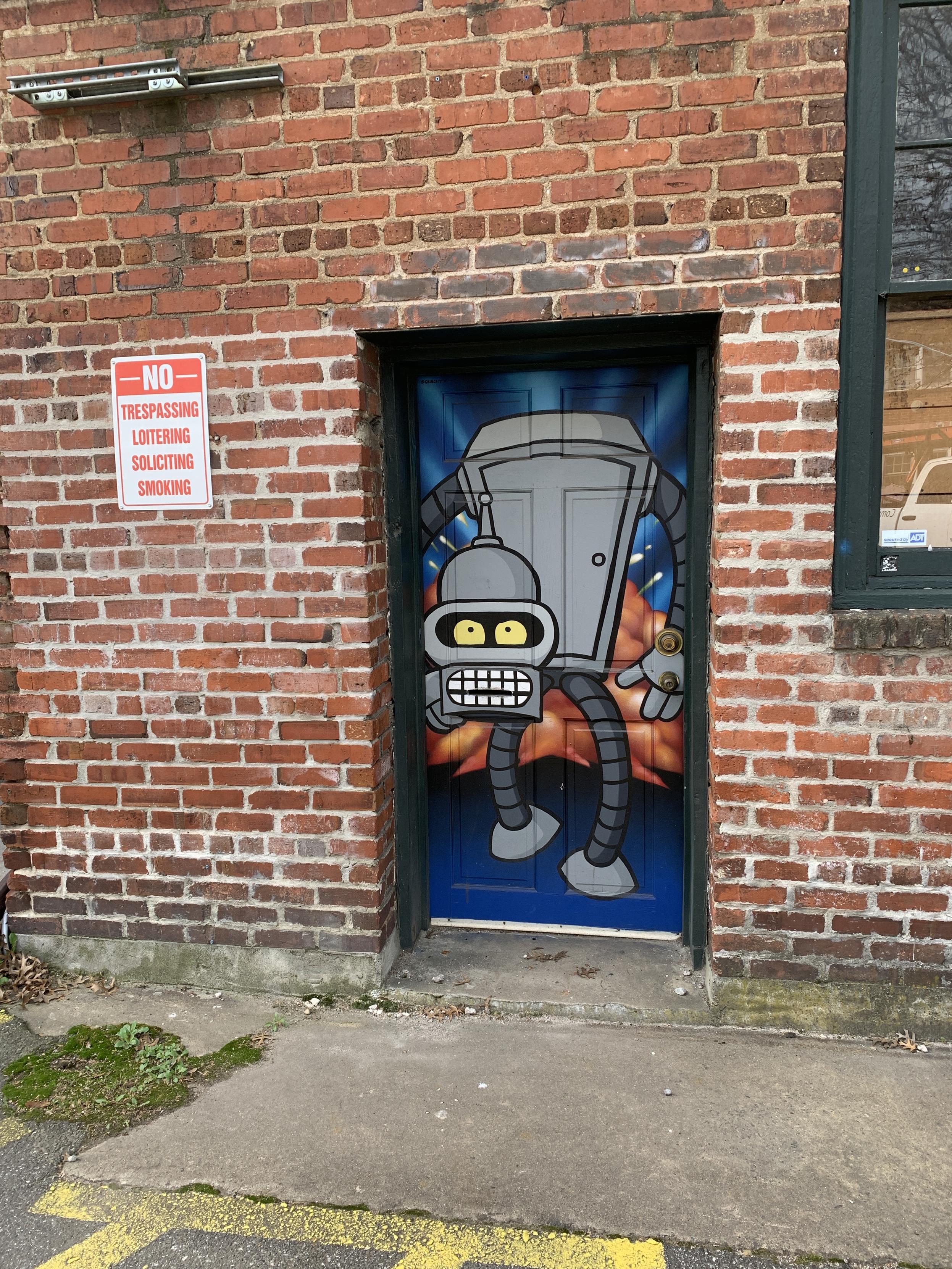 Futurama robot painted on a fire door in a brick building.  He is carrying his own head, which has quite a rictus of a grin.  Never seen the program, so no idea of the context, sorry.

This is I believe the backdoor of a comic/graphic art shop in Asheville.