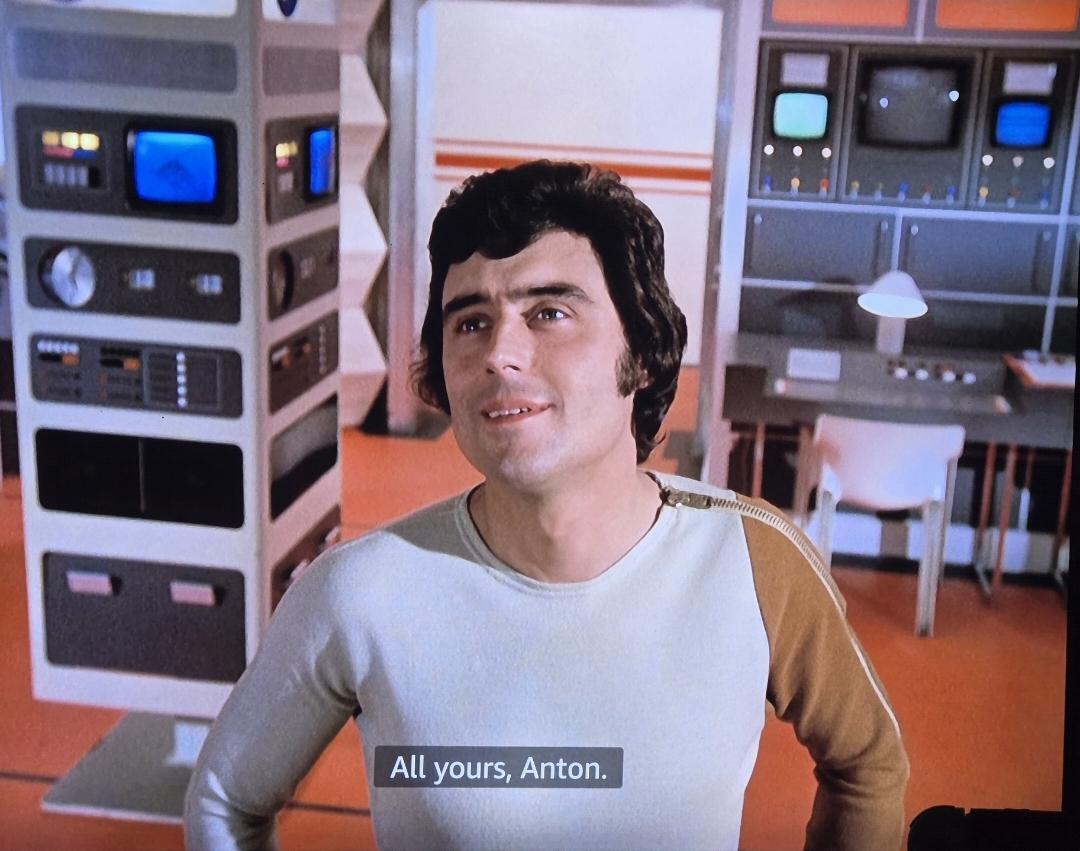 A man that looks like a young Ian M Shane in one of the rooms in the spaceship of Space 1999. Subtitles read: all yours, Anton. 
Computer panels, TV screens, desk, lamp, all very futuristic behind him.