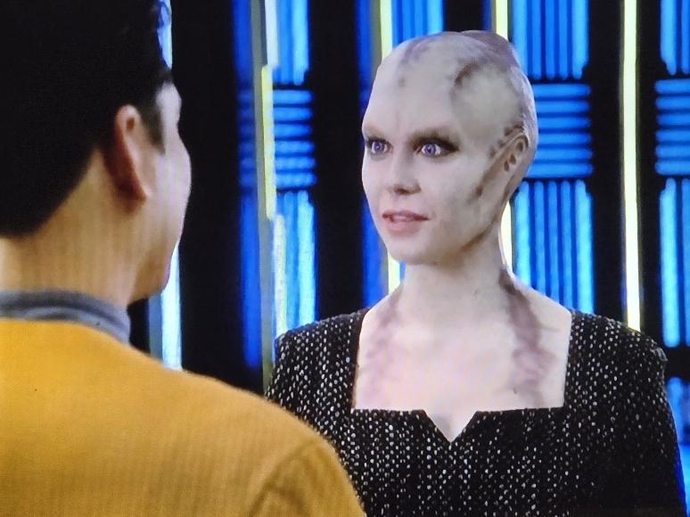 A Kobali (humanoid alien with no hair, scalp ridges and ears flush against their heads and grey markings around their neck and chest) stands by the transporter while saying goodbye to Ensign Kim.
