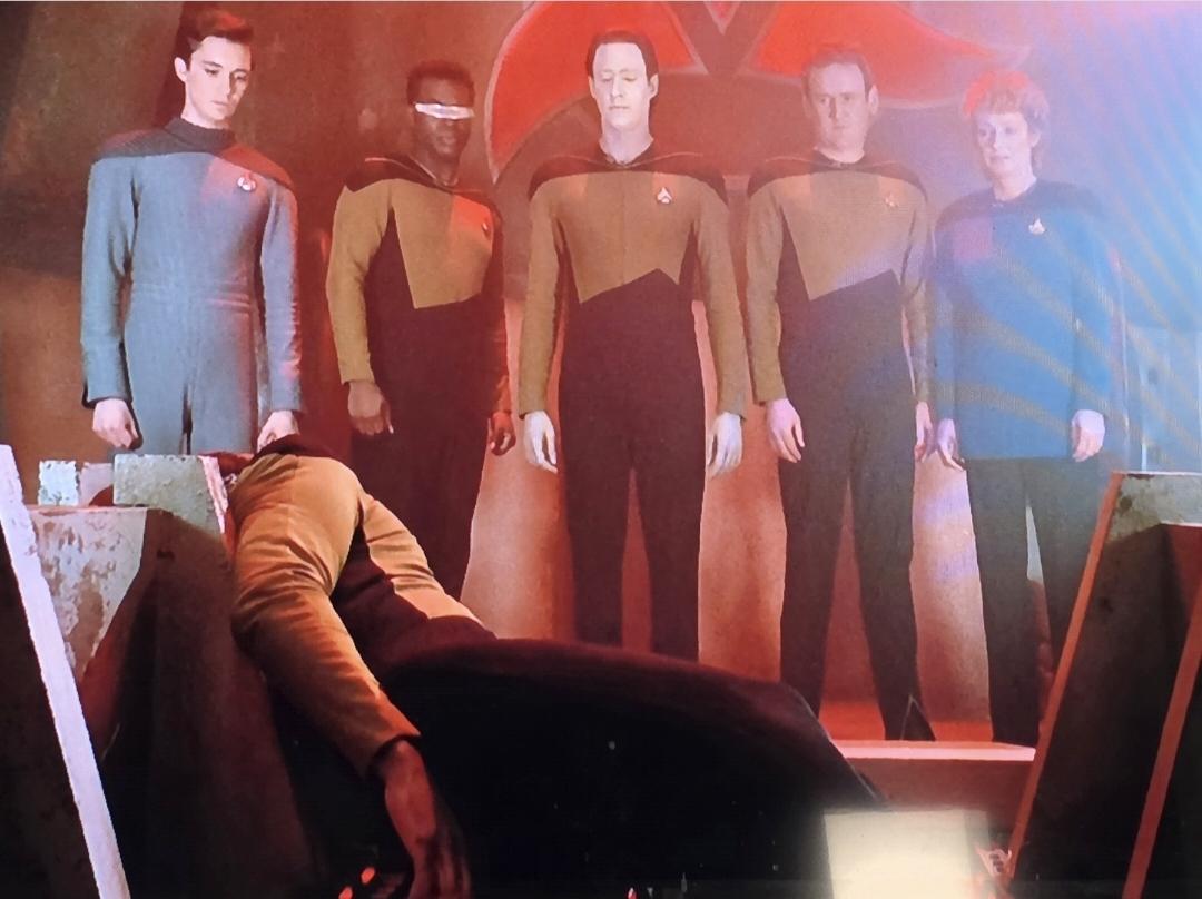 Worf on the ground having finished a reenactment of the Klingon Rite of Ascension. Looking down at him are Wesley, Geordi, Data, O'Brien and Dr. . Behind them is the red Klingon symbol.