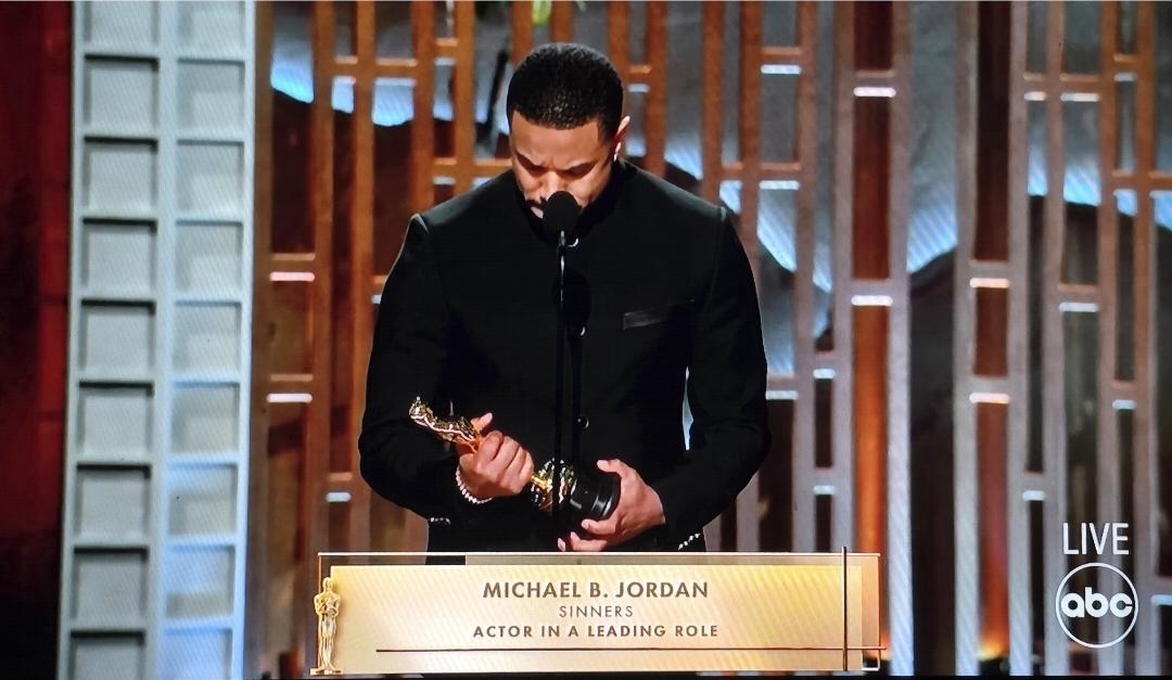 Michael B Jordan receives a standing ovation from his peers and co-nominees for his #Oscars Best Actor win. He stands at the podium looking down at the statuette while he collected his thoughts. He is wearing a very handsome modern collarless black tuxedo.