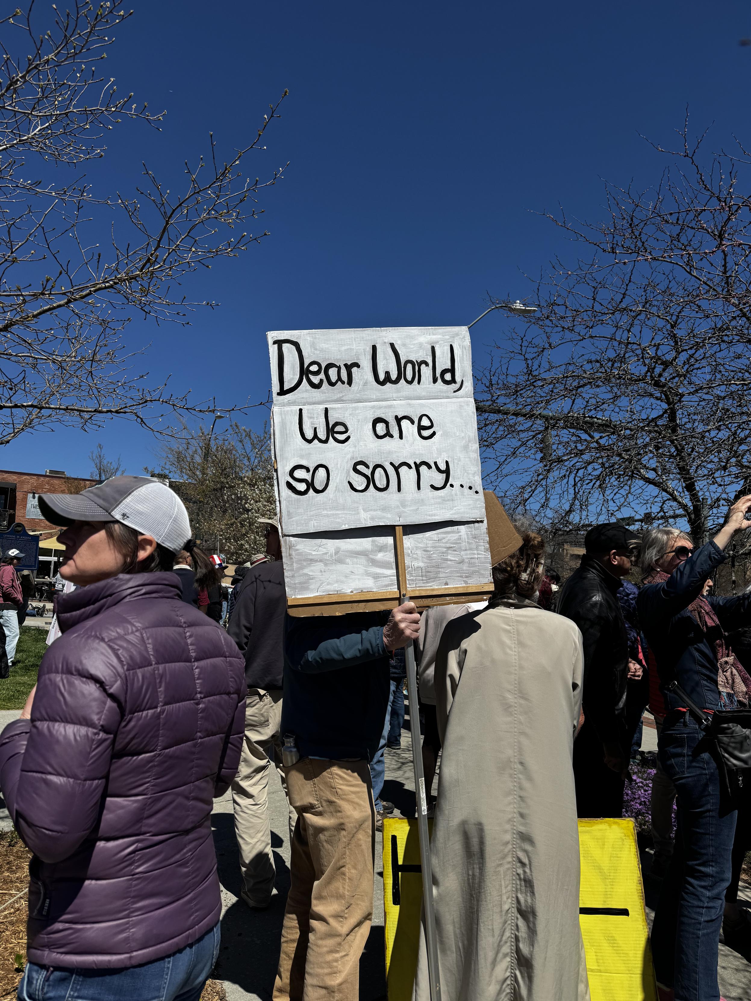 The back of several people in the crowd, one man holding up a sign which reads "Dear World, we are so sorry."