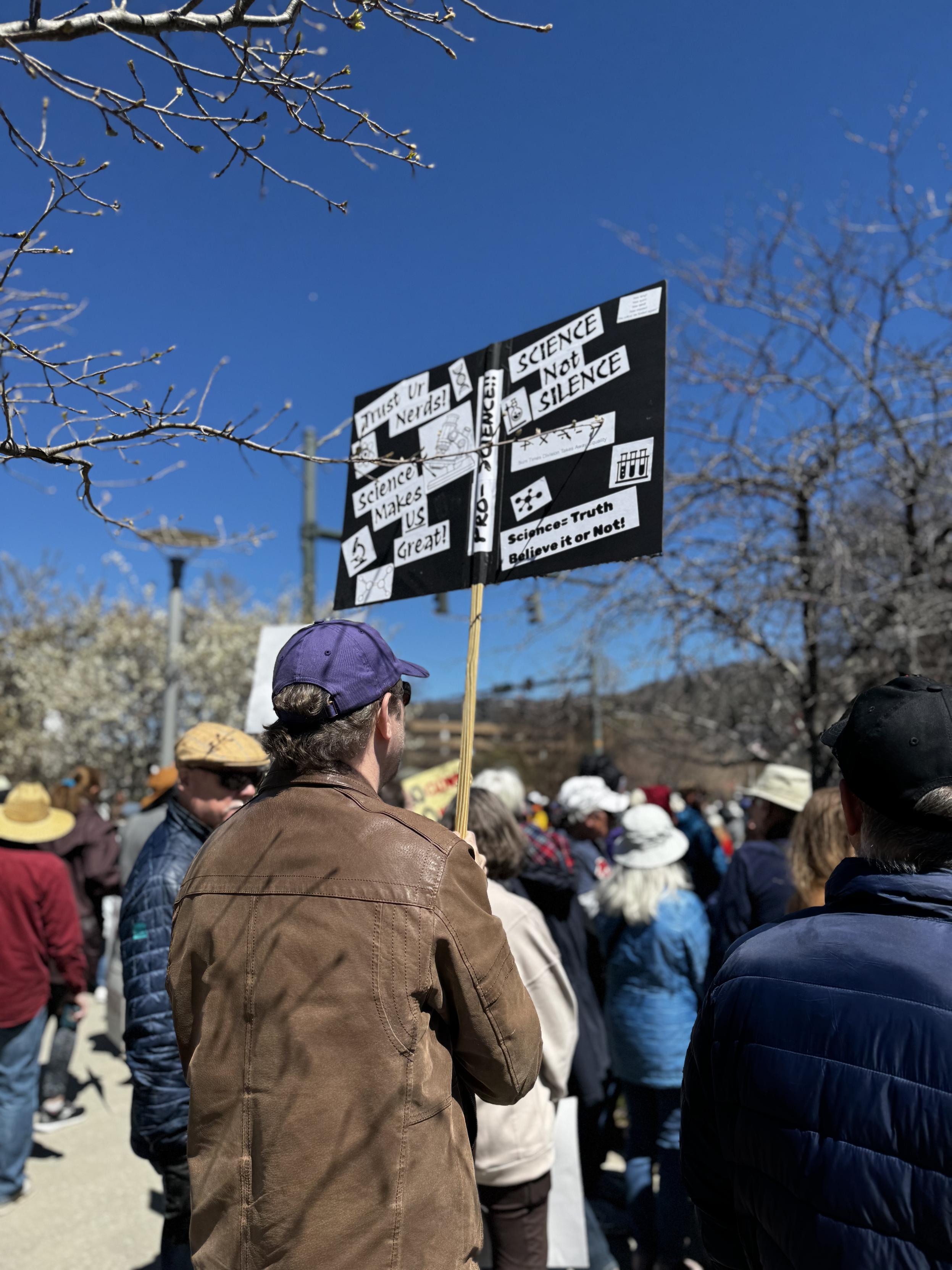 Again from behind, a man is holding up a sign with many statements about science, including "Science not silence" and "Trust Us Nerds"

Amen.