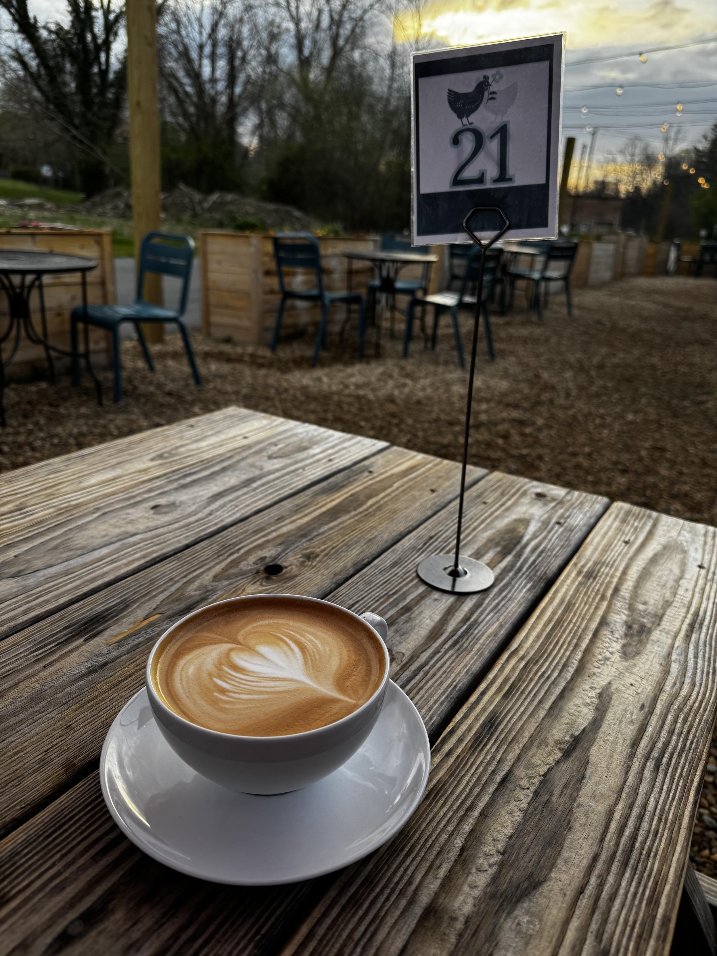 A latte with heart foam art sits on a wooden picnic table. A small sign with a chicken and the number 21 is also there for when our breakfast sandwich is delivered. A very nice walk and a great start to the day