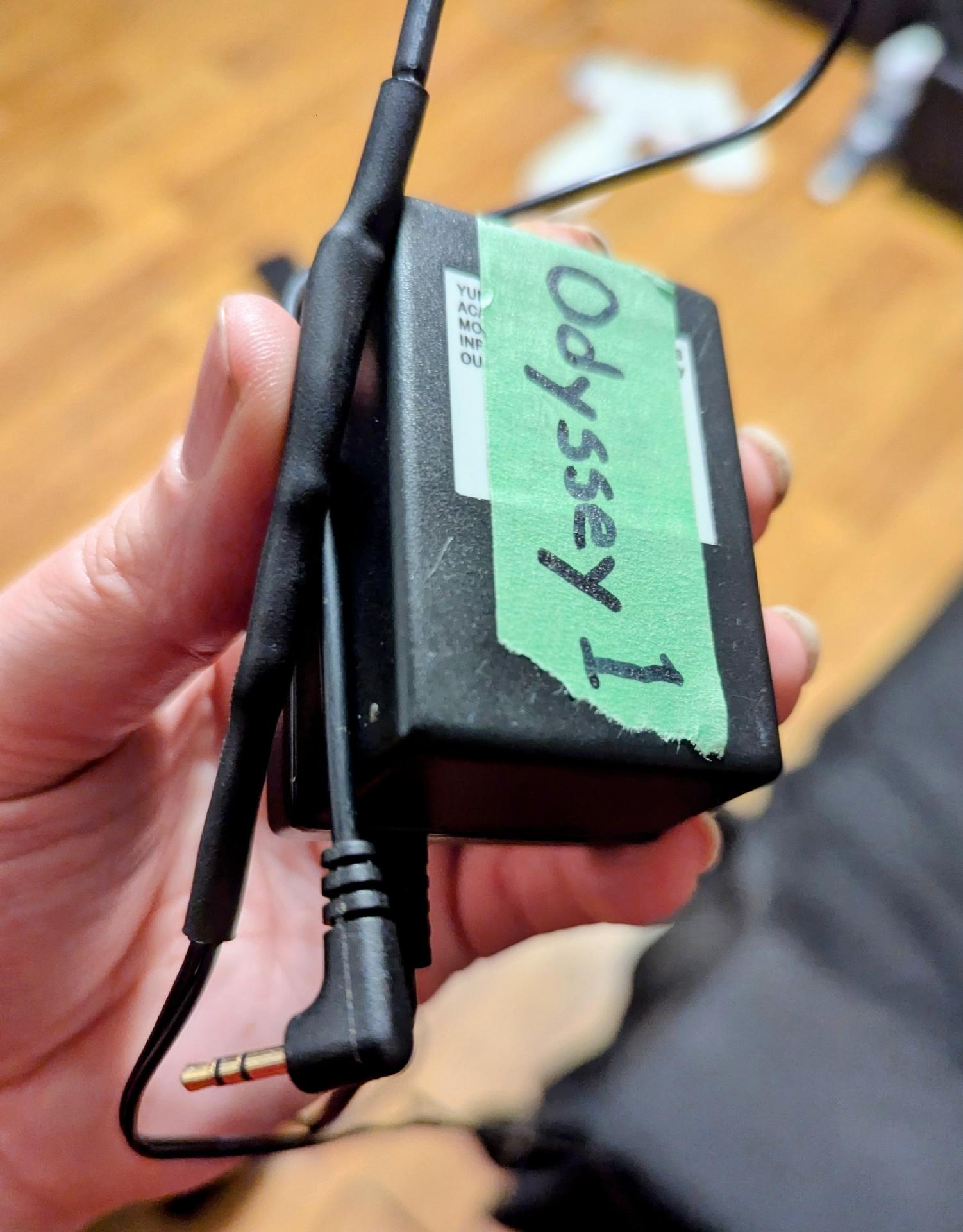 My hand holding a power adapter showing heat shrink tubing on the cable to a miniplug, painter's tape on the adapter says "Odyssey 1" written in hand with a marker