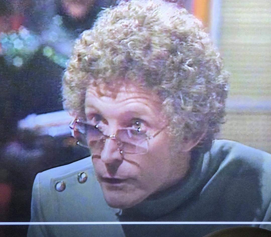 A man with curly light brown hair very shiny gold sunglasses and a mod seafoam suit looks over his eyeglasses.