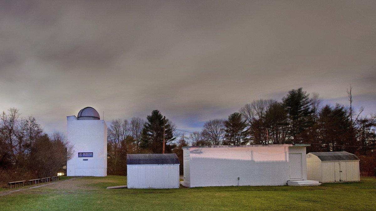 White observatory tower with a dome next to several small white utility buildings under a cloudy sky surrounded by trees.