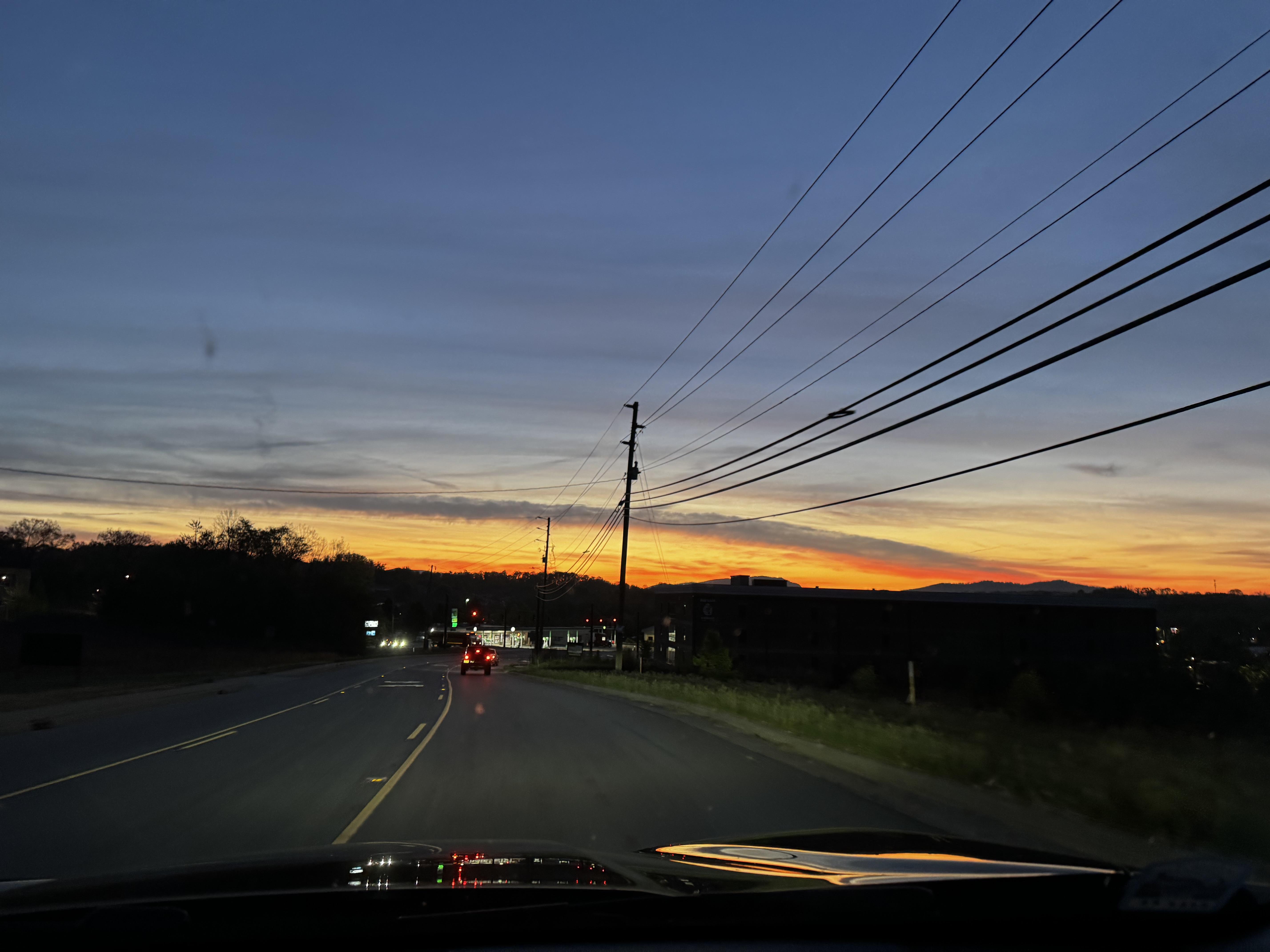 An attractive sunrise across half the sky this morning, if one can ignore the poles, wires and various important businesses.

A dark landscape (of a road and ancillary stores) is topped by a lightening blue sky and low rows of orange and yellow.  About 6:40 this morning, so actual dawn is not far off.