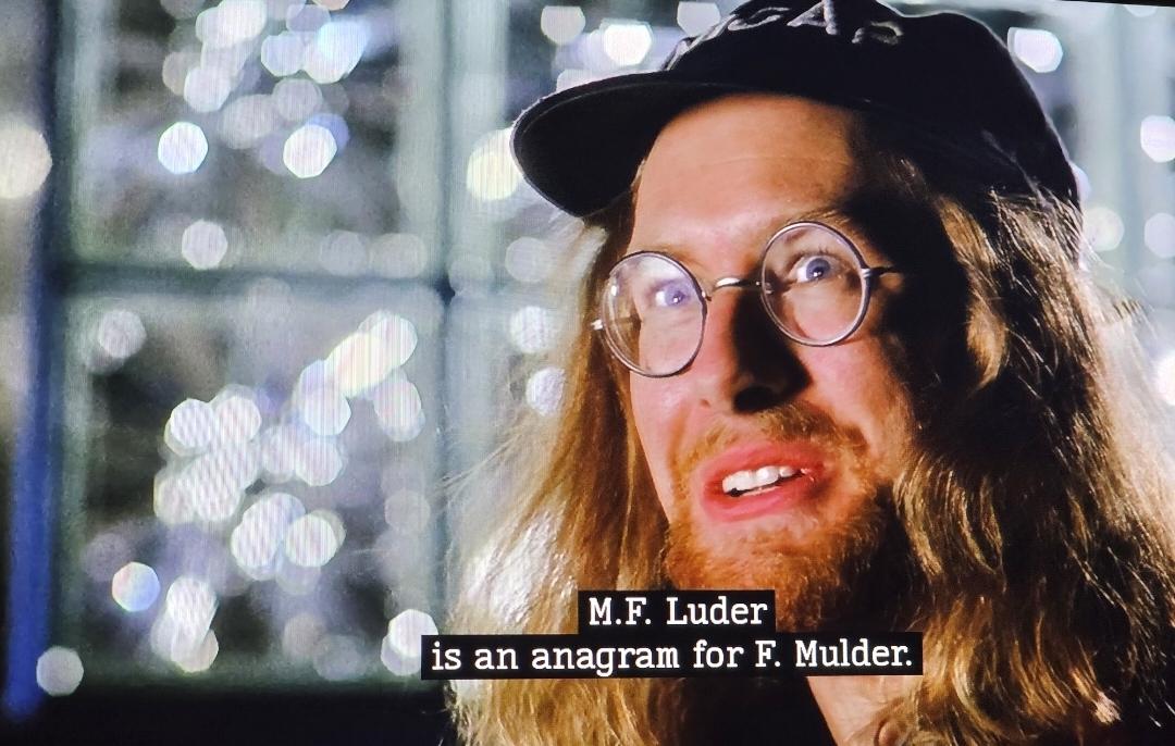Scott Below as Max Fenig on X-Files. He is a man in his 30s with round wireframes glasses long red hair and beard sporting a NICAP black cap. He smiles as he tells Mulder, "M.F. Luder is an anagram for F. Mulder."