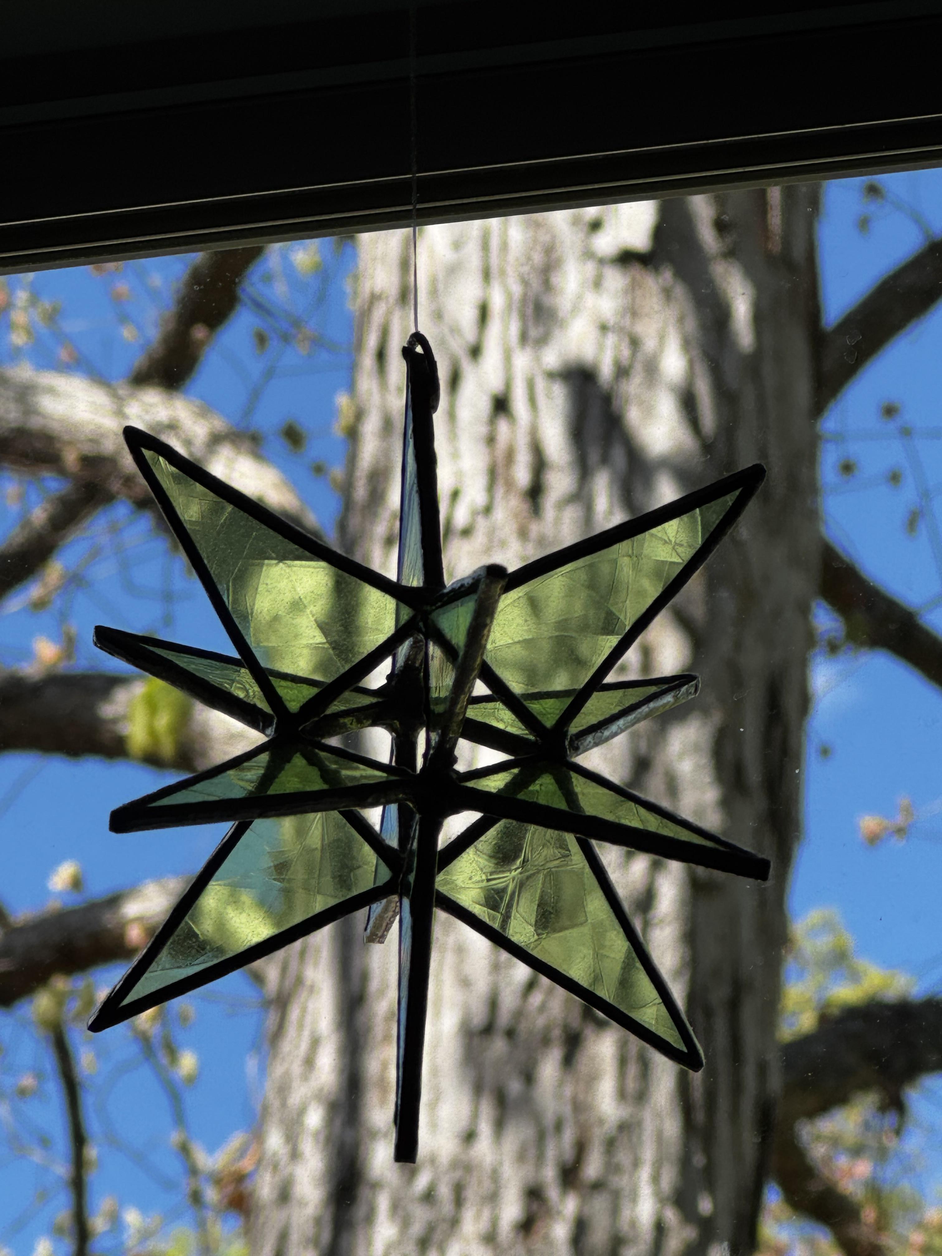 An object comprised of 12 triangular pieces of green glass hangs in a window.  It is made of 3 planes of 4 triangles each, so there is an open square area in the center.  I'm sure there's a name for this shape, but I don't know it.  It's particularly welcome in this room in the winter, when the oak is not yet leafing out.

No sun is coming through at this time of day, but the trunk of the oak tree behind it allows you to see the color.