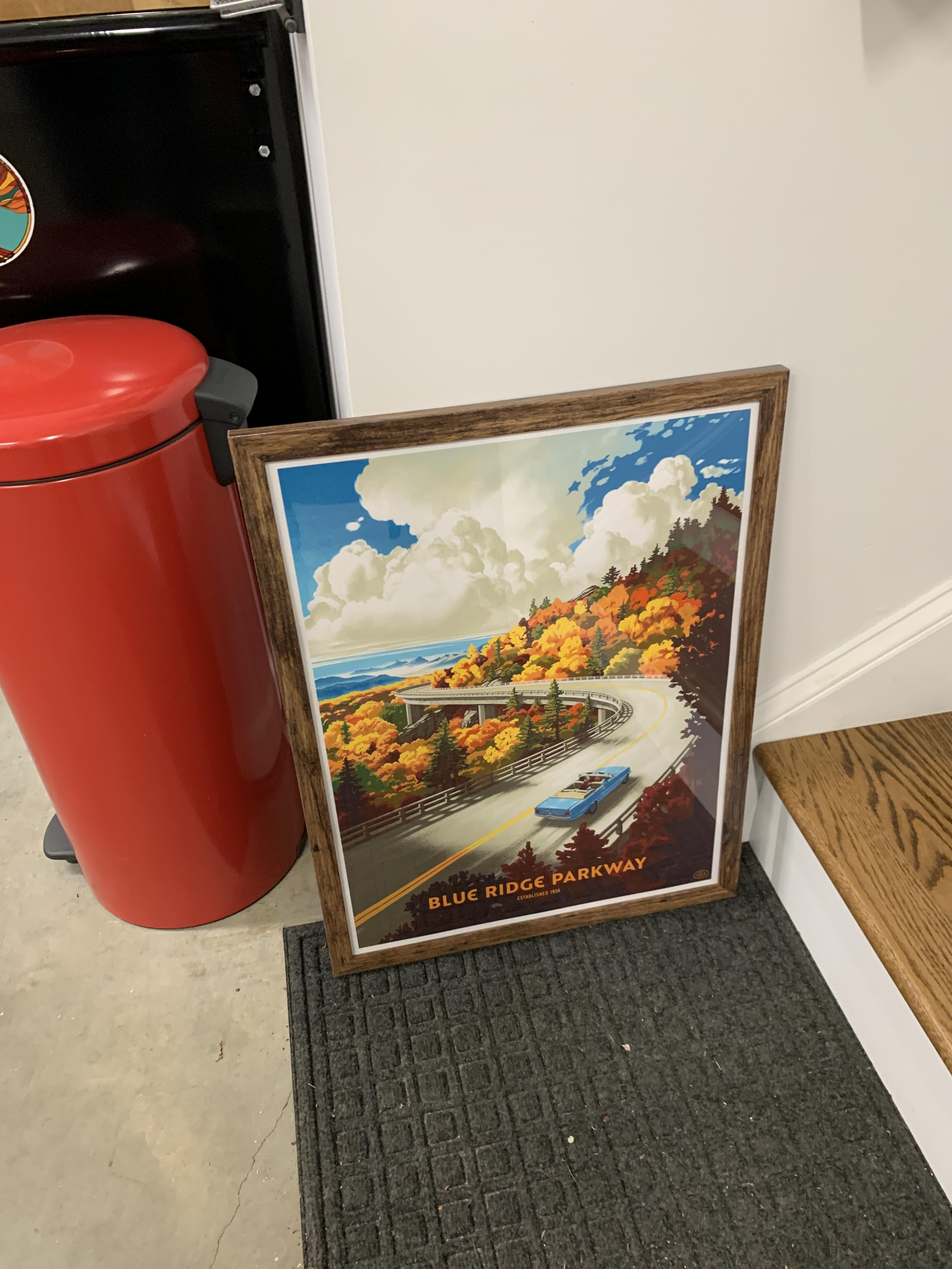 A colorful, travel-poster style art print of the BRP in fall, with the road curving through the gorgeous autumn colors, and a vintage blue convertible enjoying the drive.

The framed print is sitting on the floor near stairs and a red waste paper can, awaiting being hung up in front of my car in the garage, so the Subaru can dream of being out on the road even when it is parked.