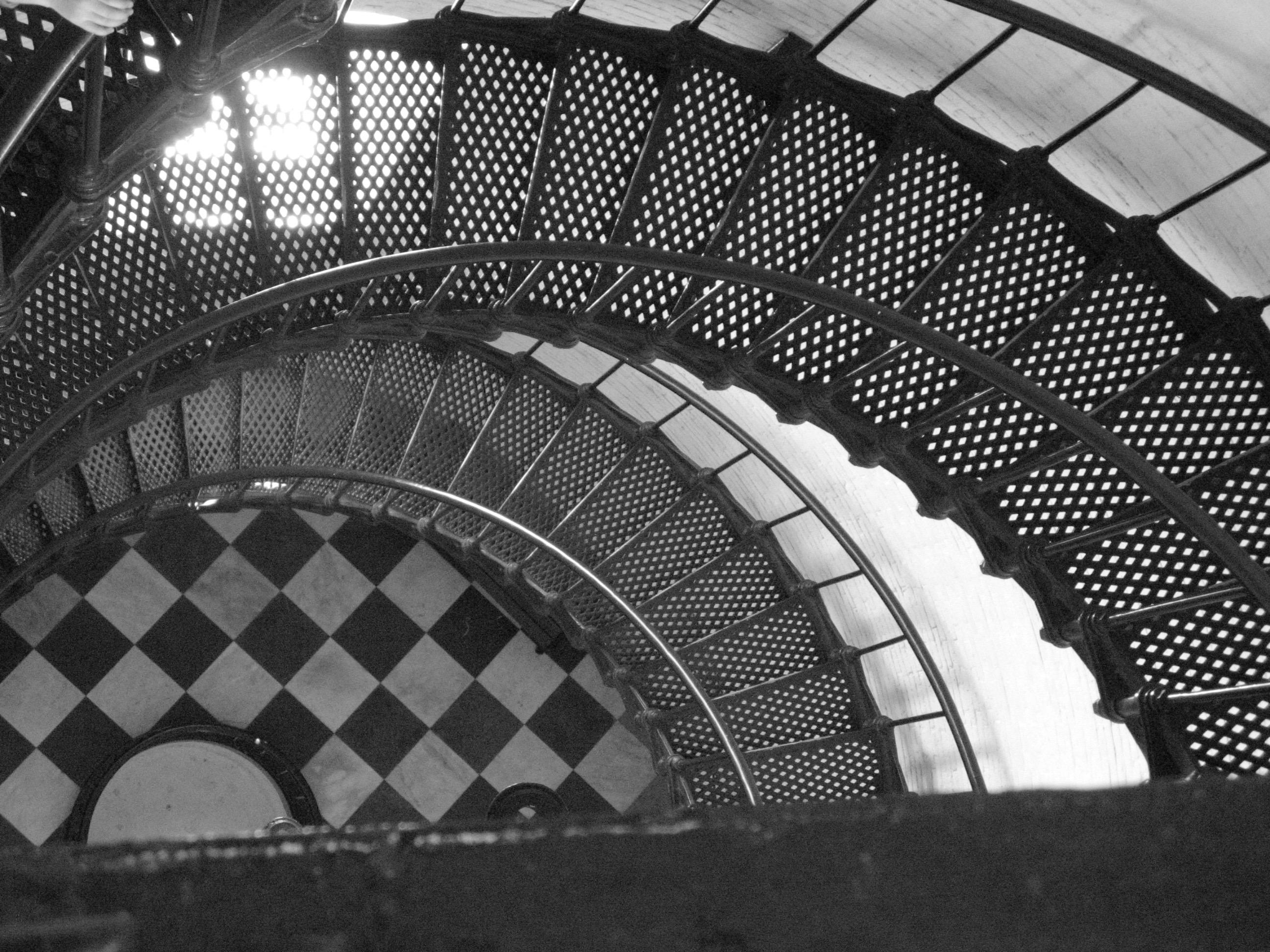 An off-center black and white (I think) looking down at the checkerboard floor at the bottom of the metal mesh spiral staircase in the lighthouse.  The photo includes about 1/4 of the spiral and 2 turns of the stairs.