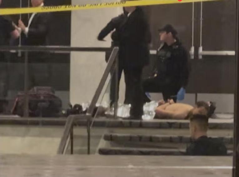 Four policemen are shown around or near the suspect who is laying face down on the ground near some doors. He is naked and handcuffed behind him. A foil blanket covers him from the waist down.