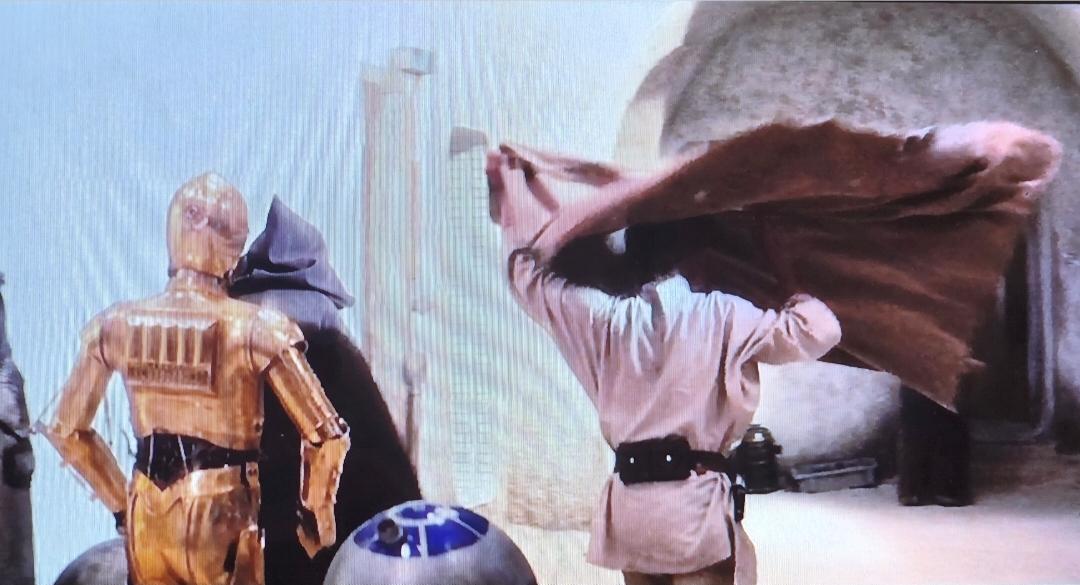 Kenobi, R2D2 C3PO and Luke are walking outside in Mos Eisley. Luke tosses his poncho in the air to get it over his head.