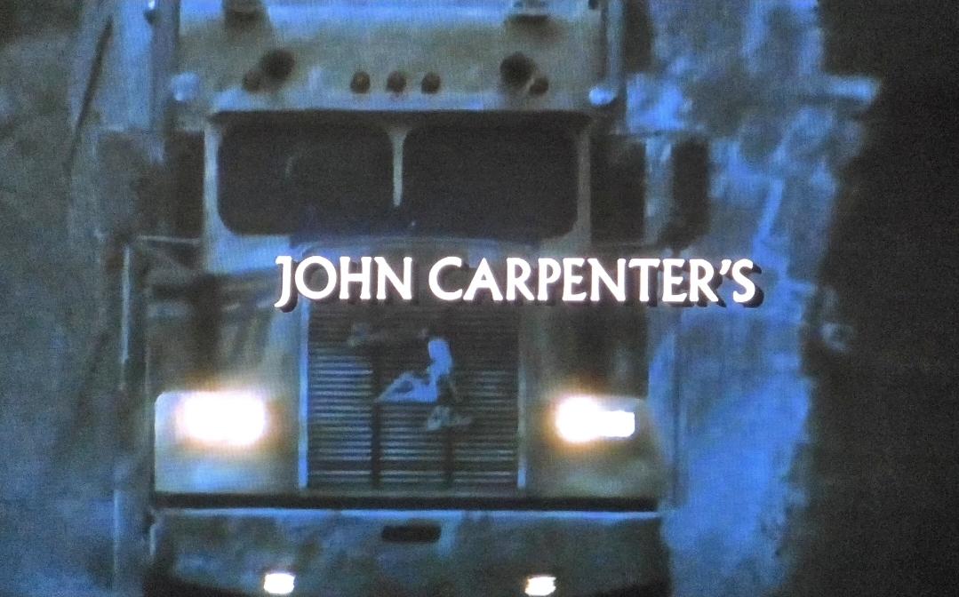A semi drives in the evening on a rural road. There's a silhouette of a sexy woman in the truck grille. The road looks wet.
Superimposed credits read: John Carpenter's