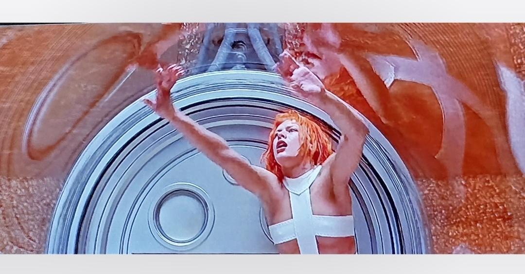 Milla Jovovich as Leeloo inside the clear chamber she was reconstructed in. It's tubular in shape and she's touching the top and looks confused/distressed. She has short red hair and is wearing a bandage strap across her breasts.