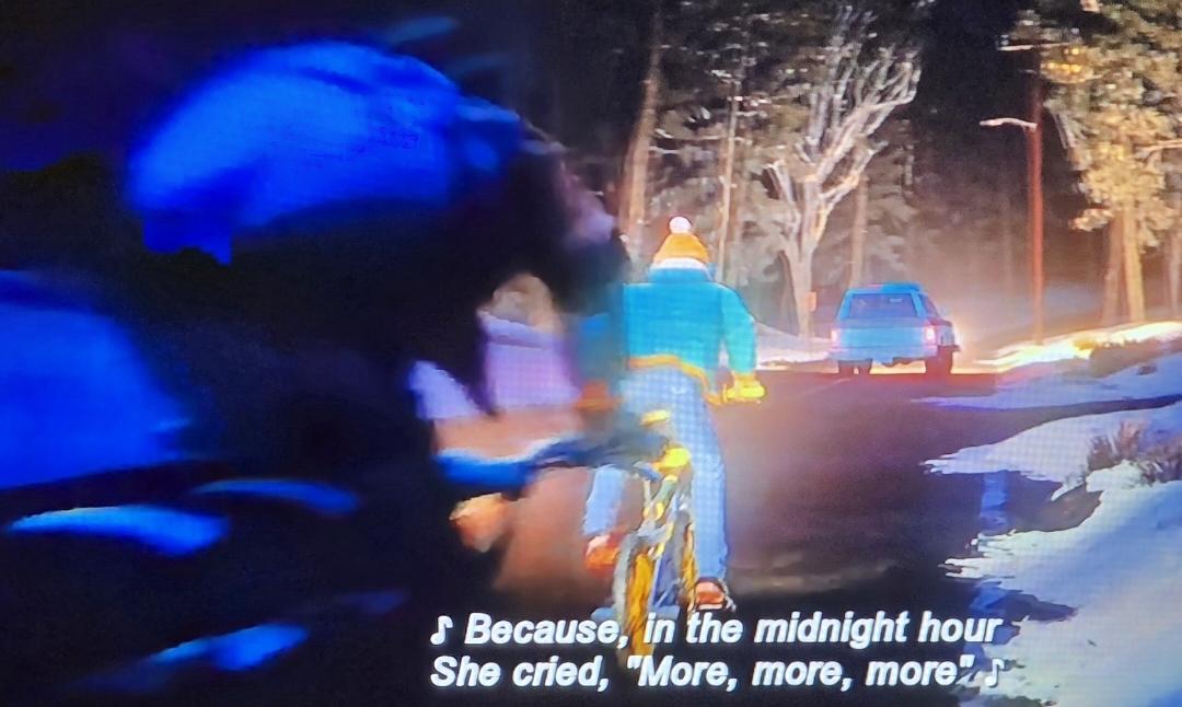 Two kids are racing on their bikes down a country winter road at night. Ahead of them we see Hopper's truck. The music playing is Billy Idol's Rebel Yell. "Because in the midnight hour she cried, 'More, more, more'"