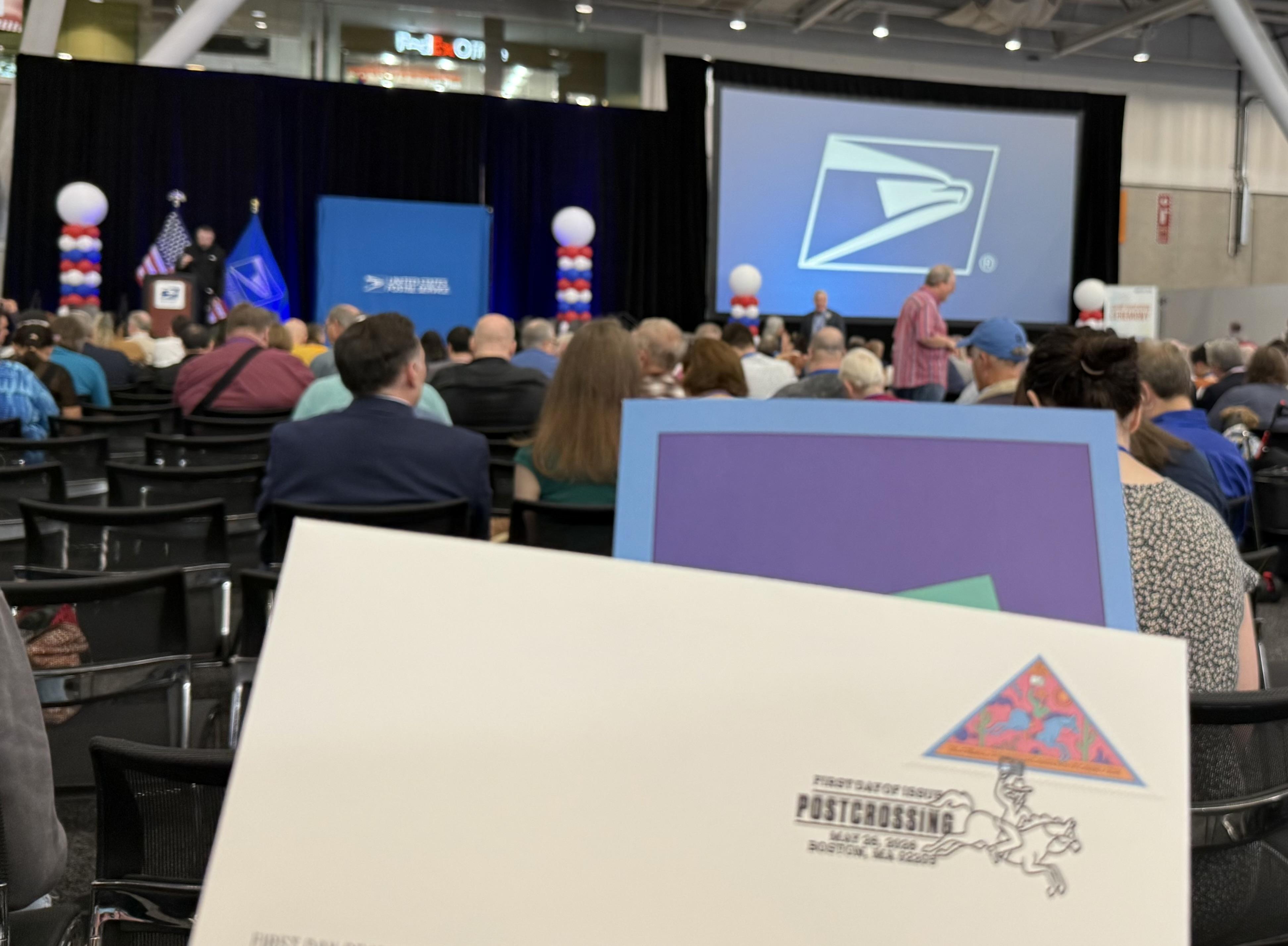 Photo of the stage with the USPS logo, I’m holding up a first day of issue cancellation card. Lots of folks in the background