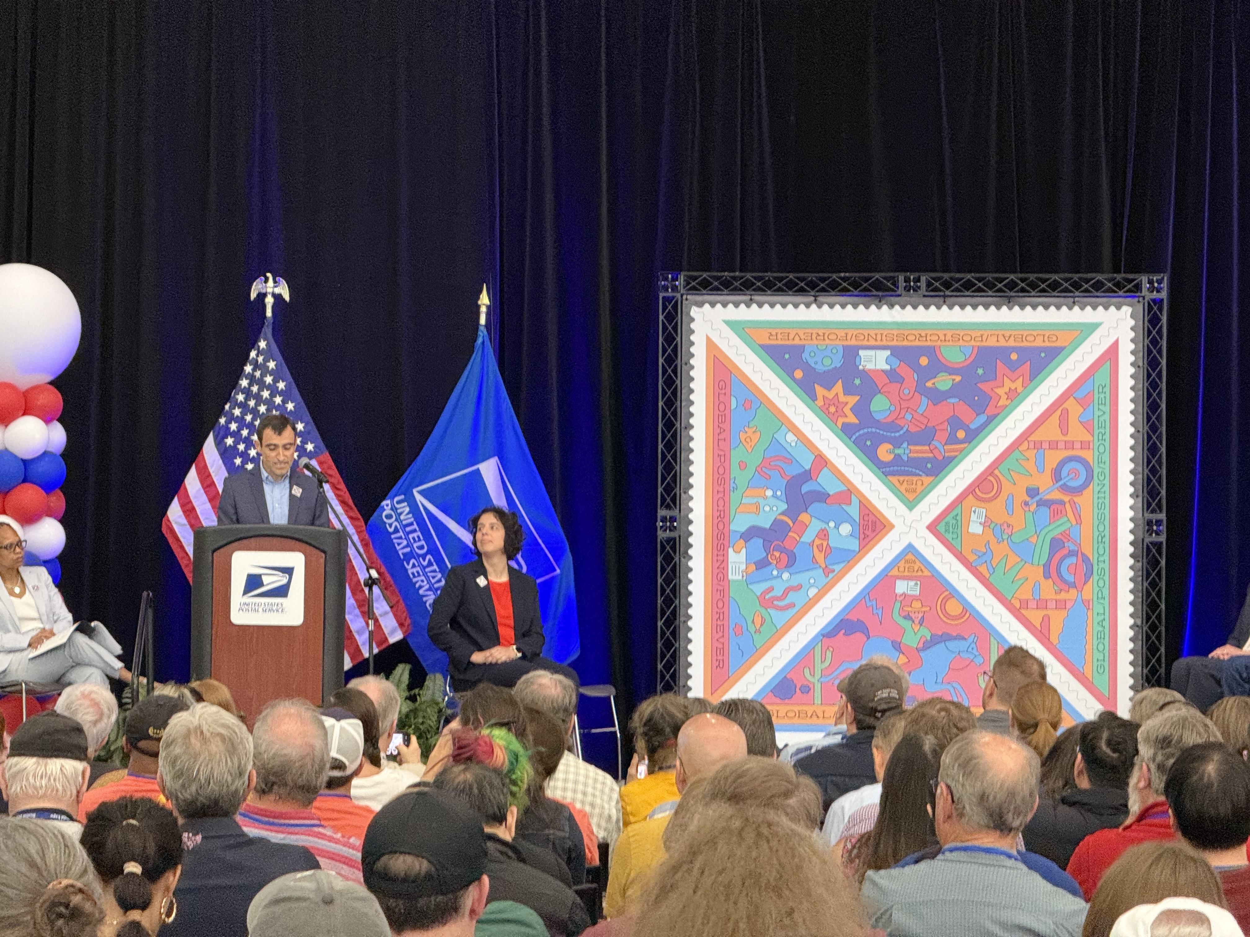 Slim fellow with dark hair in a blue suit behind a USPS podium flanked by giant replicas of the new postcrossing triangle stamps