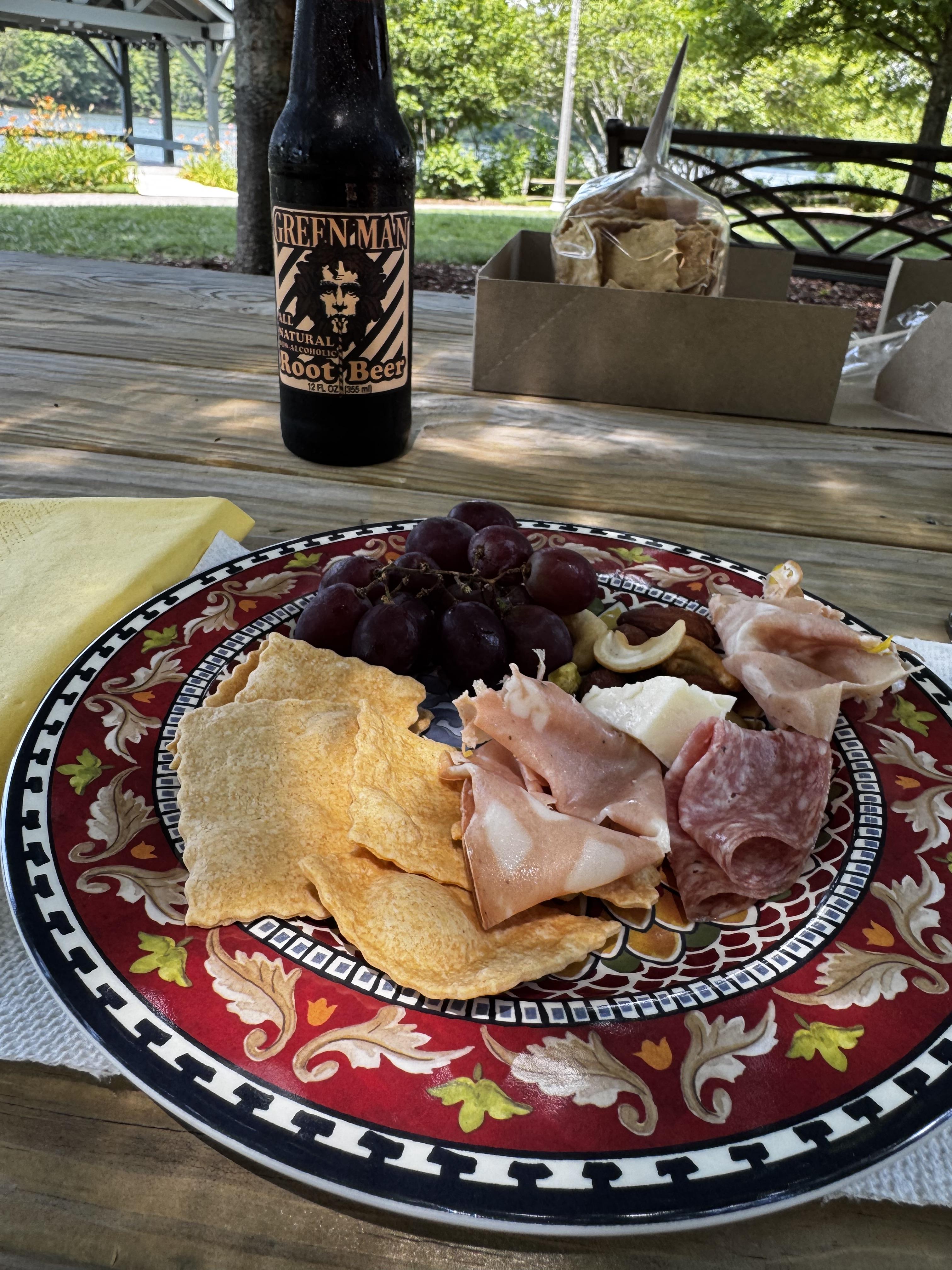 A decorative plastic plate sits on a wooden picnic table, layered with various cold cuts, nuts, grapes, olives and really excellent Root and Branch crackers.  The bottle of root beer is behind the plate, with the lake somewhat visible in the distance.

Green Man is, of course, known for its beer, and it took me a while before I noticed that their eponymous totem on this label is drinking out of a striped straw.  While the root beer was okay, for the price I would go for one of their dark beers instead.