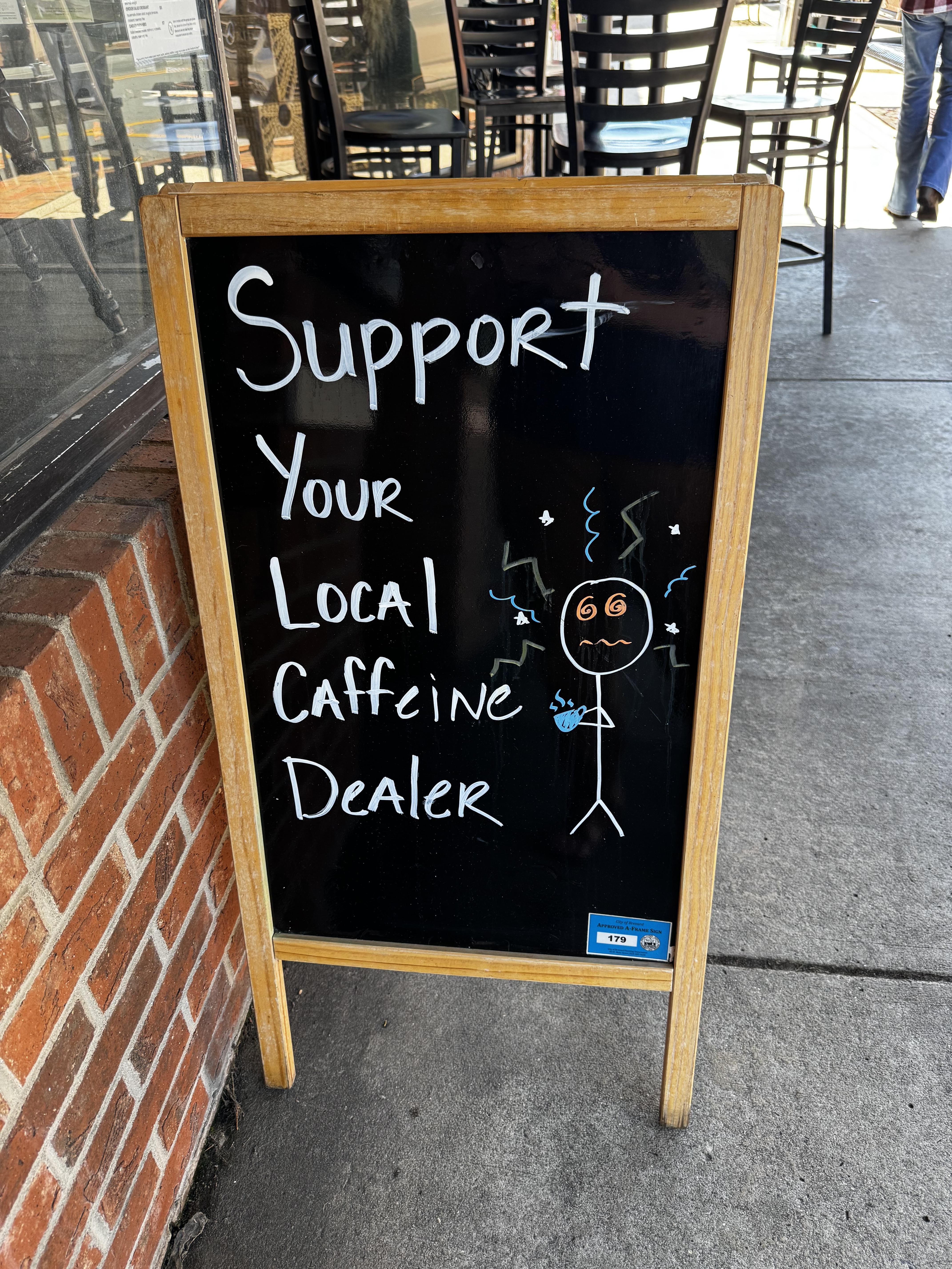 A wooden A-frame chalkboard is placed outside a coffee shop on a sidewalk.  It has a drawing of a stick-figure human, with hurricane eyes, and lightning for hair, holding a cup of coffee.

The legend states:  "Support Your Local Caffeine Dealer"